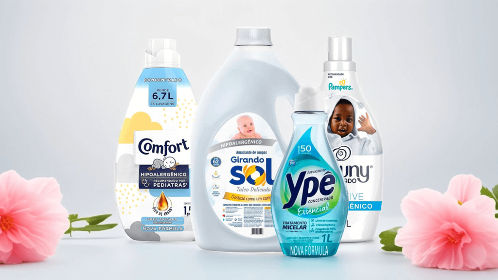 Imagem com embalagens das principais marcas de amaciantes hipoalergênicos, incluindo Comfort, Girando Sol, Ypê e Downy, destacadas em um fundo claro com flores, representando suavidade, maciez e cuidado para peles sensíveis.